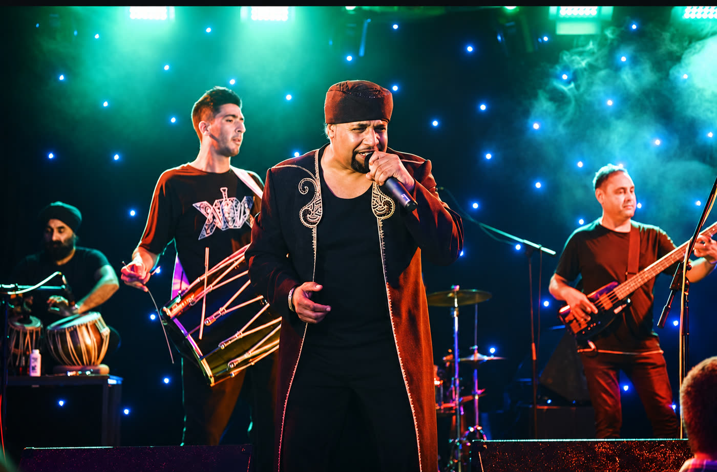 Achanak live: singer in an embroidered coat with dhol and bass players under a starry backdrop